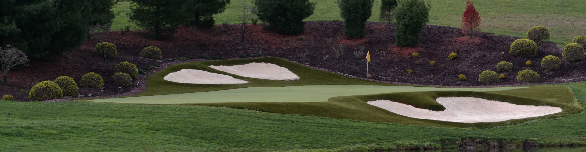 Artificial turf installation on a golf course green with a yellow flag pin, white sand bunkers, a pond water hazard, and manicured evergreen and boxwood landscaping